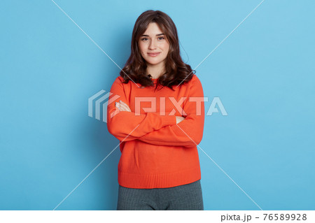 Smiling young brunette woman girl in sweater posing isolated blue background, alluring lady holding hands crossed, looks at camera with satisfied and happy expression. Smiling young brunette woman girl in sweater posing isolated blue background, alluring lady holding hands crossed, looks at camera with satisfied and happy expression. 76589928