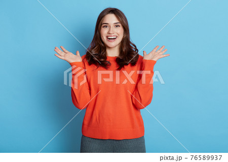 Happy pleased European woman keeps hands spreading aside, focused at camera, notices desirable things, laughs, dressed in orange sweater, poses against blue background. Happy pleased European woman keeps hands spreading aside, focused at camera, notices desirable things, laughs, dressed in orange sweater, poses against blue background. 76589937