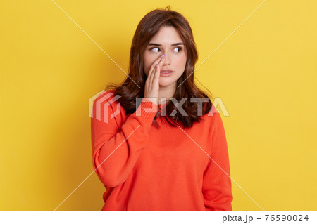 Mysterious dark haired woman keeps palm near mouth, looks aside and tells secret or private information, lady wearing in orange jumper gossips about something, stands indoor against yellow wall. Mysterious dark haired woman keeps palm near mouth, looks aside and tells secret or private information, lady wearing in orange jumper gossips about something, stands indoor against yellow wall. 76590024