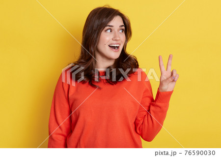 Portrait of joyful smiling pretty girl wearing orange jumper, showing v sign with fingers and looking aside with excited facial expression, shows peace or victory gesture, rejoicing success. Portrait of joyful smiling pretty girl wearing orange jumper, showing v sign with fingers and looking aside with excited facial expression, shows peace or victory gesture, rejoicing success. 76590030