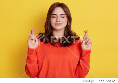 Pretty superstitious woman crosses fingers, prays for better, looks calm and relaxing, closes eyes, wears orange jumper, poses indoor over bright yellow background, anticipates important results. 76590032