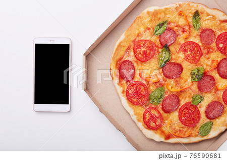 Pizza in box and smartphone with blank screen on white table, top view of delicious pepperoni isolated over light background. Modern mobile phone near box with pizza. Copy space for advertisment. Pizza in box and smartphone with blank screen on white table, top view of delicious pepperoni isolated over light background. Modern mobile phone near box with pizza. Copy space for advertisment. 76590681