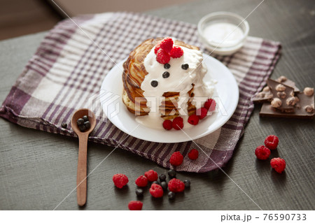 Picture of stack of pancakes with blueberries and raspberries on white plate, dark brown colour r backdrop decorated with cup of tea, spoon, pieces of chocholate and berries. Delicious breakfast. 76590733