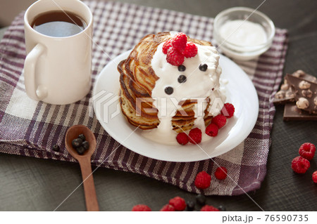 image of home made pancakes with sour cream on white plate decorated with berries, fresh blueberries and raspberries, cup of tea or coffee, spoon, pieces of chocholate on cotton checkered towel. 76590735