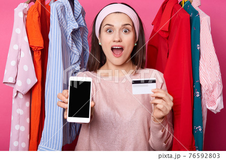 Portrait of excited young dark haired girl shows plastic credit card which full of money while holding mobile phone isolated over rose background. Smiling female shows phone, woman gets salary. Portrait of excited young dark haired girl shows plastic credit card which full of money while holding mobile phone isolated over rose background. Smiling female shows phone, woman gets salary. 76592803