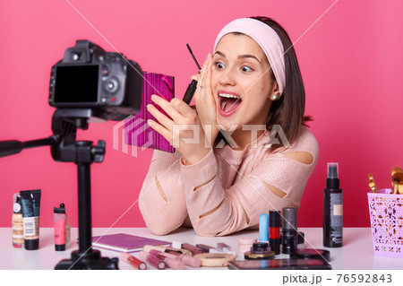 Image of woman or beauty blogger with eye shadow brush, mirror and camera, sitting at table, surrounded with cosmetics, recording makeup tutorial video at home. Blogging, technology and beauty concept 76592843