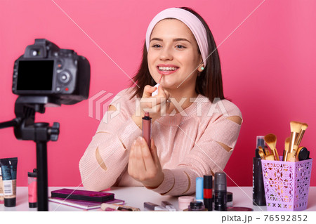 Attractive charismatic young female applying lipstick, demonstrating quality of product in front of camera, smiling. Positive magnetic black haired model wearing light pink sweater and headband. 76592852