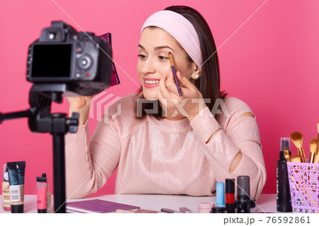 Beautiful female records video. Yong blogger shows how to apply eyeshadow. Lady advetises cosmetics on her channel. Woman holds mirror and brush against pink wall. Fasion and blogging concept. 76592861