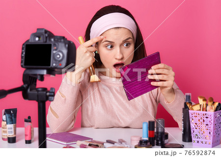 Close up portrait of surprised young woman beauty vlogger, looks at mirror and shows it in her blog, charming girl sits at desk in front of camera, sits with opend eyes and astobished expression. 76592874