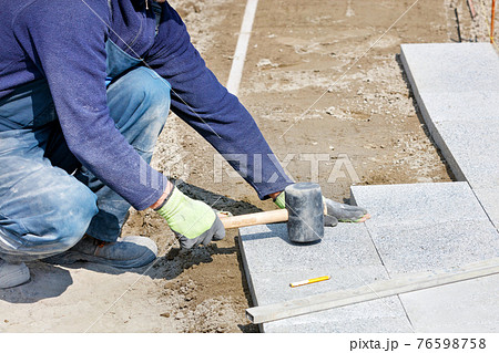 A worker places granite blocks on the sidewalk with a rubber hammer on a marked area. A worker places granite blocks on the sidewalk with a rubber hammer on a marked area. 76598758