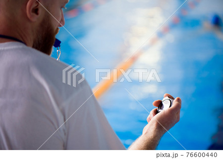 A young swimming coach with a stopwatch in his hand watches the training session near the sports 76600440