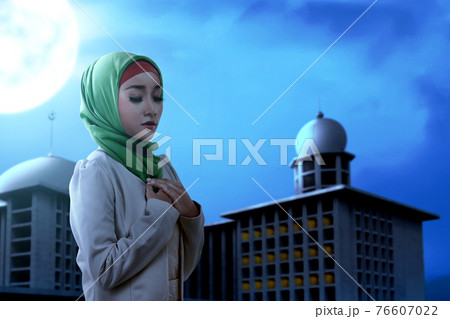 Asian Muslim woman in a veil standing while raised hands and praying 76607022