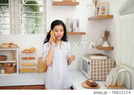 Portrait of smiling young beautiful woman standing, drinking water and talking on smartphone in kitchen. Portrait of smiling young beautiful woman standing, drinking water and talking on smartphone in kitchen. 76615174