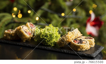 Custom sushi roll in tempura with nori, fresh salmon, tuna, avocado, masago caviar, drizzled with pineapple sauce with salad pouring as decoration on a black plate on a wooden table for Christmas. 76615500