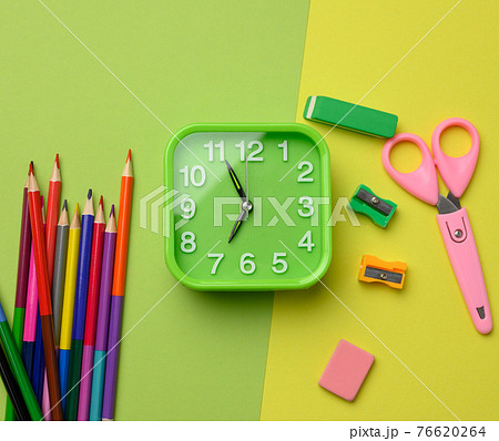 square green clock and stack of multicolored wooden pencils on a green background, top view 76620264