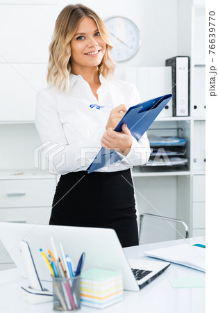 Well-wishing girl with folder of documents is standing in well-lit office Well-wishing girl with folder of documents is standing in well-lit office 76639770