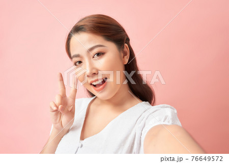 Happy expressive young woman posing while taking pictures of herself through cellphone, over pink background 76649572