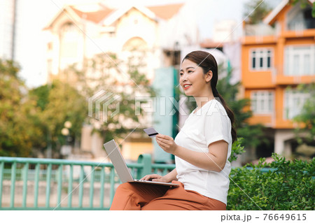 Happy young woman or girl sitting in the park with laptop and bank credit card and smiling after successful online shopping 76649615