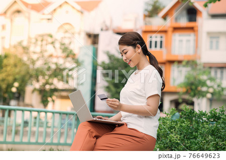 Photo of happy asian woman using laptop and holding credit card while sitting on bench in green park 76649623