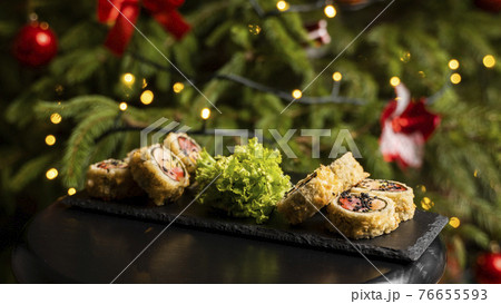 Custom sushi roll in tempura with nori, fresh salmon, tuna, avocado, masago caviar, drizzled with pineapple sauce with salad pouring as decoration on a black plate on a wooden table for Christmas. 76655593