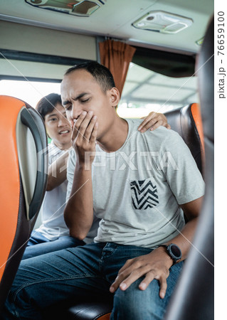 a travel sick young man and his friend massage his back while sitting on a bus bench 76659100