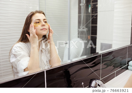 Caucasian woman applying hydrogel patches on under eye area, looking at mirror. Happy young lady enjoying morning anti age anti wrinkle routine in bathroom Caucasian woman applying hydrogel patches on under eye area, looking at mirror. Happy young lady enjoying morning anti age anti wrinkle routine in bathroom 76663921