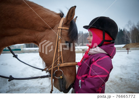 Close up of a little girl jockey smiling and looking into the horse eyes, in the profile. Girl horse rider with a horse in a winter open riding hall, looking eye to eye. Girl and horse friendship, equ Close up of a little girl jockey smiling and looking into the horse eyes, in the profile. Girl horse rider with a horse in a winter open riding hall, looking eye to eye. Girl and horse friendship, equ 76665772