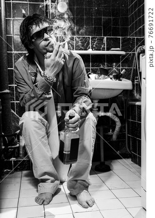 Drunk Man sits in a toilet with a bottle of whiskey and cigarette 76691722