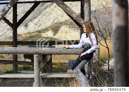 A girl sits on a wooden fence near an old gazebo made of wooden beams on a sunny warm day A girl sits on a wooden fence near an old gazebo made of wooden beams on a sunny warm day 76693470