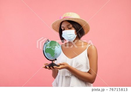 Travel ban during covid lockdown. Sad black woman in summer dress wearing face mask and holding globe on pink background 76715813