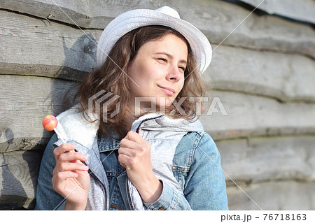 Portrait of dreamy girl with a lollipop near wooden fence. Beautiful young caucasian woman with sweet Chupa-Chups. Spring holidays in ferme. 76718163