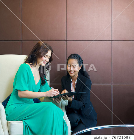 The hotel manager in black suit hands a receipt to a caucasian customer. A young tourist sign the bill paying for a hotel stay while sitting on a cream armchair. The hotel manager in black suit hands a receipt to a caucasian customer. A young tourist sign the bill paying for a hotel stay while sitting on a cream armchair. 76721098