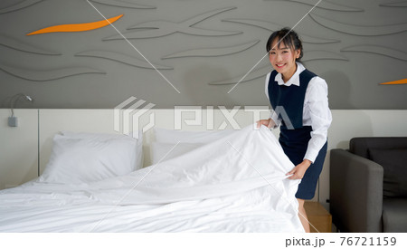 Young hotel maid with a smile making the bed. Staff in blue uniform preparing room for hotel guest. 76721159