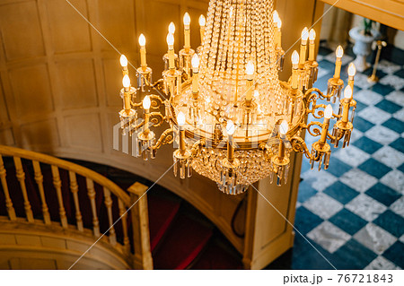 Top view of an antique chandelier. A huge crystal gold chandelier with candles, against the backdrop of a spiral staircase and a checkerboard floor. Top view of an antique chandelier. A huge crystal gold chandelier with candles, against the backdrop of a spiral staircase and a checkerboard floor. 76721843