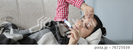 Woman with cold lying on sofa under covers and looking at thermometer Woman with cold lying on sofa under covers and looking at thermometer 76725039