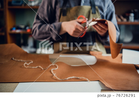A young tanner in his workshop at the table cuts out leather elements of shoes A young tanner in his workshop at the table cuts out leather elements of shoes 76726291