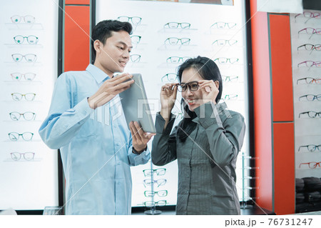 a shop assistant holding a tablet while a female customer tries on the glasses to look at the tablet screen while standing 76731247