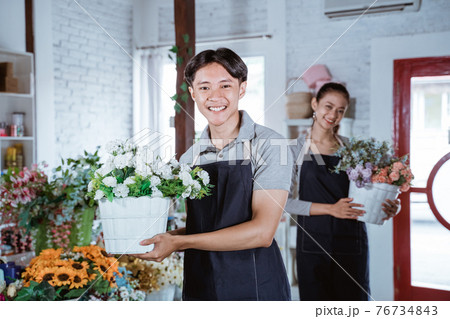 happy young man florist wearing apron holding bucket flower smiling looking at camera. working in flower shop happy young man florist wearing apron holding bucket flower smiling looking at camera. working in flower shop 76734843