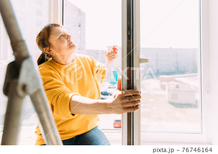 Woman Of Fifty In Yellow Sweater And Jeans Washes Dusty Window In Apartment. 50 Year Old Woman Cleans Windows From Stains Using Rag And Spray Cleaner. Caucasian Elderly Woman Is Cleaning House, Doing 76746164