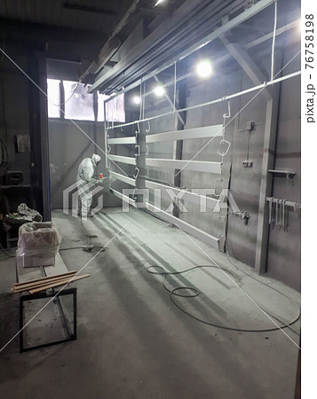 A male painter in a white disposable protective suit paints a metal structure with a spray gun at a paint factory 76758198