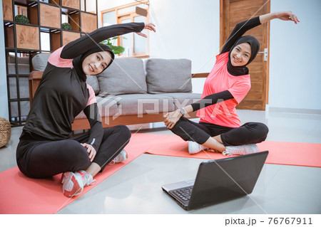 two women wearing hijab sportswear sit cross-legged on the floor with their bodies leaning to the side and hands up while warming up two women wearing hijab sportswear sit cross-legged on the floor with their bodies leaning to the side and hands up while warming up 76767911