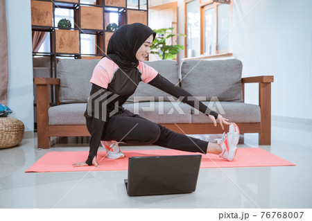 a woman wearing a hijab gym clothes when squatting stretches with one leg pulled sideways and held with one hand when in front of a laptop a woman wearing a hijab gym clothes when squatting stretches with one leg pulled sideways and held with one hand when in front of a laptop 76768007