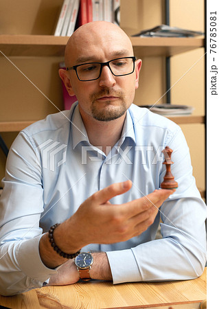bald man with glasses looking intently at chess piece. Business man holds chess queen in his hand. Looking for solution. 76785081