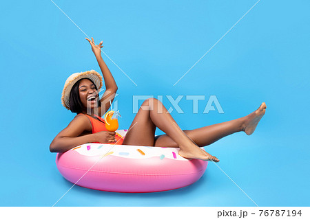 Positive black woman sitting on inflatable ring, holding tropical cocktail on blue background, full length portrait 76787194