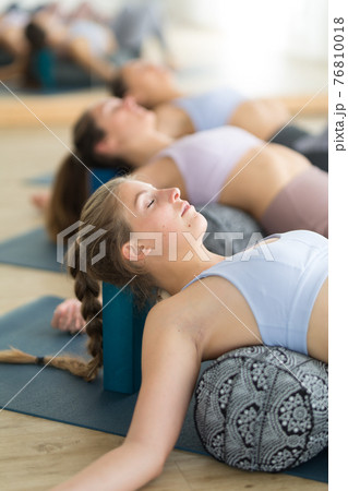 Restorative yoga with a bolster. Group of three young sporty attractive women in yoga studio, lying on bolster cushion, stretching and relaxing during restorative yoga. Healthy active lifestyle 76810018