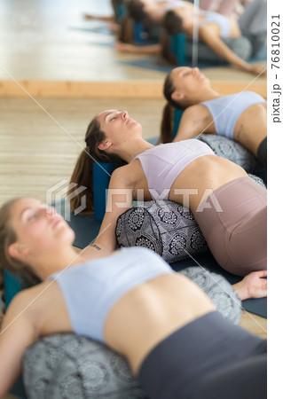 Restorative yoga with a bolster. Group of three young sporty attractive women in yoga studio, lying on bolster cushion, stretching and relaxing during restorative yoga. Healthy active lifestyle 76810021