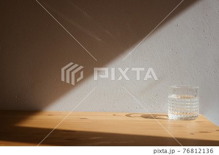 A glass with clean clear water and sharp shadows stands on a wood table on gray wall background 76812136