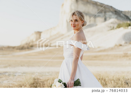 Beautiful bride in a wedding dress with a bouquet standing against white rock 76814033