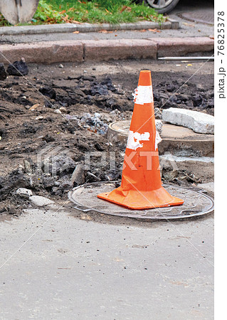 An orange traffic cone stands on the manhole and encloses the danger zone on the roadway. An orange traffic cone stands on the manhole and encloses the danger zone on the roadway. 76825378