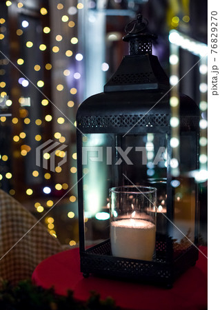 an iron lantern with glass candles and a flame with glowing warm light on a night terrace with bokeh from festive garlands on the window in defocus. 76829270